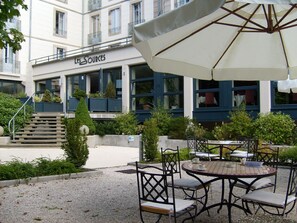 An outdoor seating area with tables and chairs, a covered umbrella, and a building with a sign reading "Les Sources".