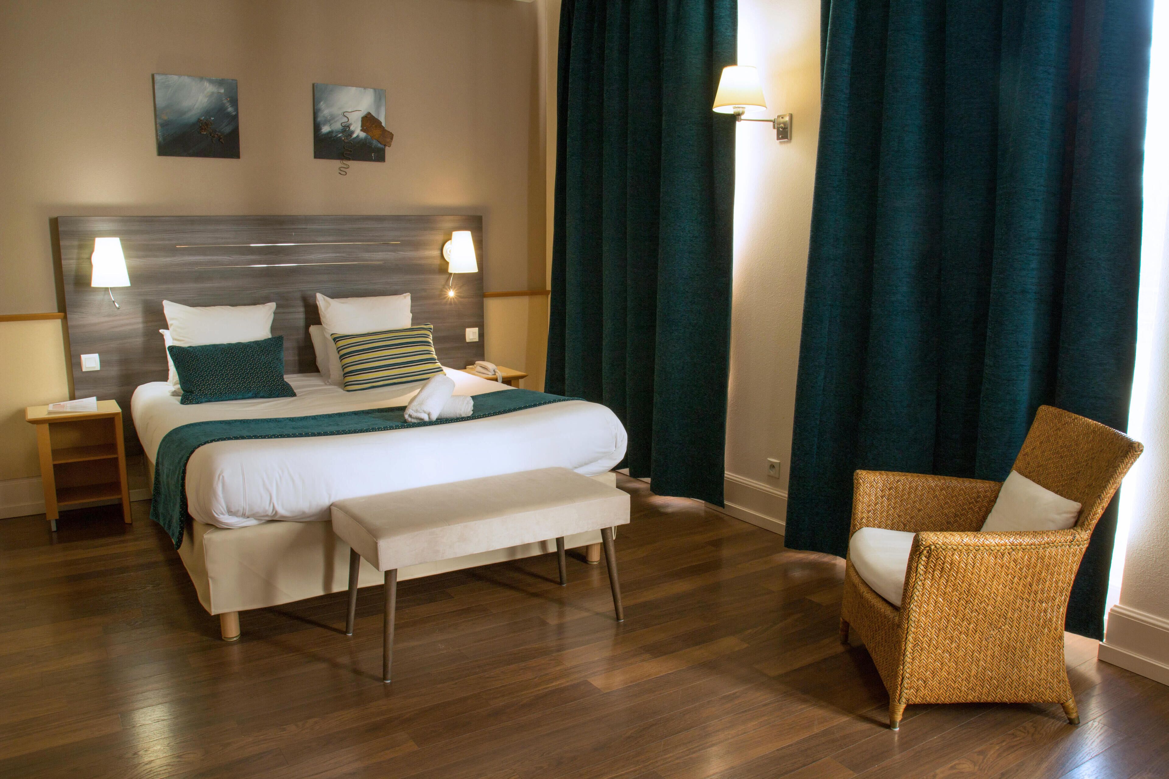 Une chambre d’hôtel avec un grand lit, une banquette, un fauteuil en osier et deux lampes fixées au mur.