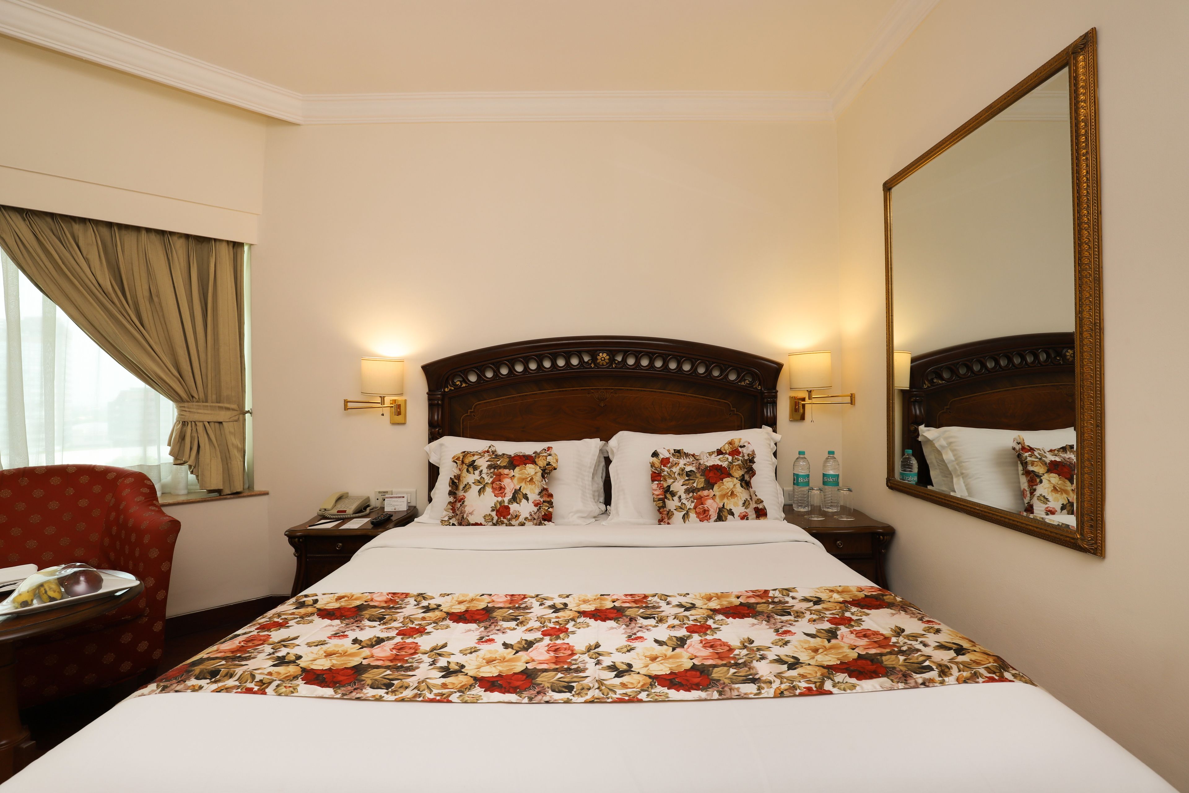 Een hotelkamer met een groot bed, een bloemenprint beddeken, twee bedlampjes, een nachtkastje met een telefoon en een spiegel aan de muur.