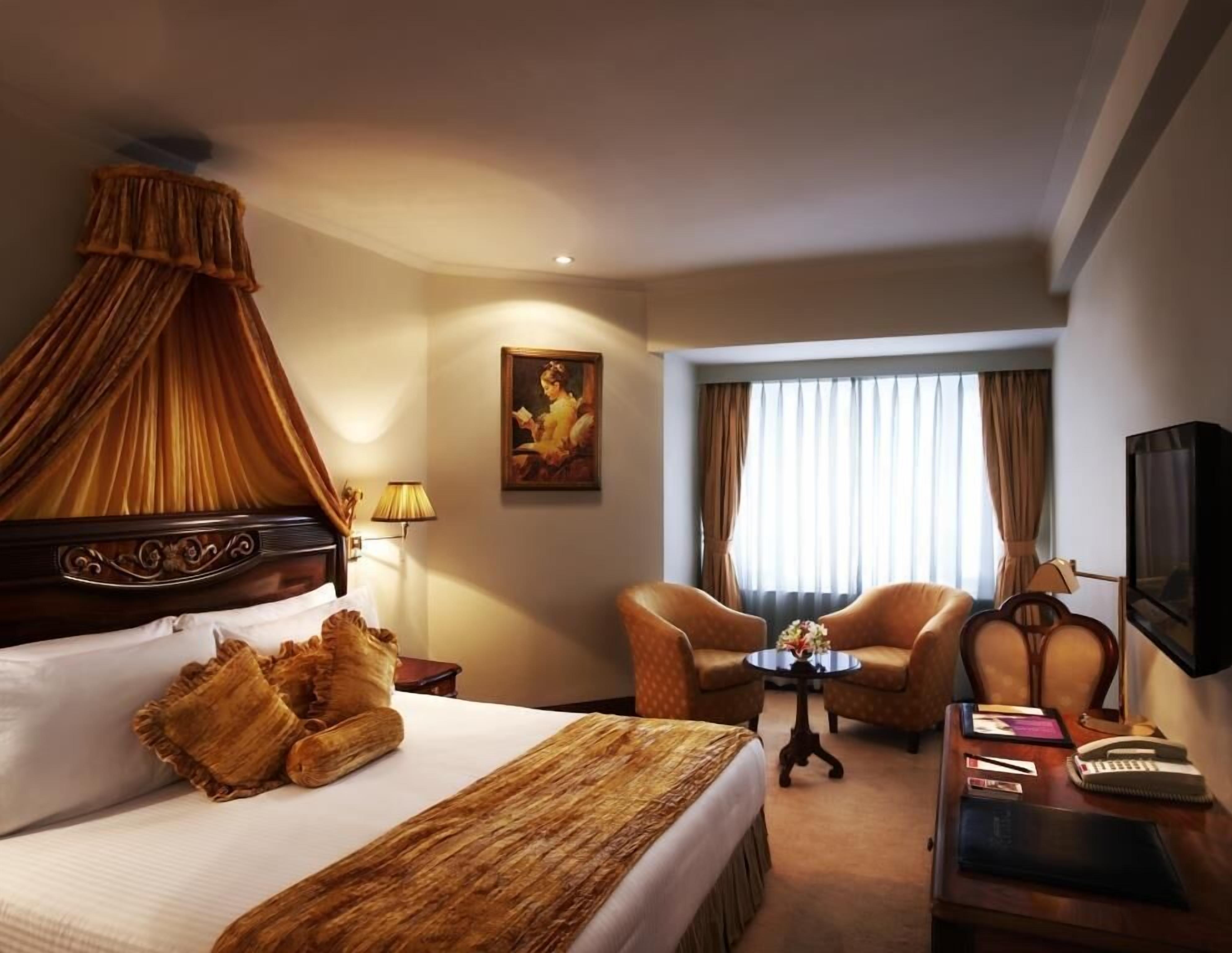 A hotel room with a bed, two armchairs, a small table, a desk with a keyboard, a television, and a painting on the wall.