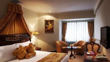 A hotel room with a bed, two armchairs, a small table, a desk with a keyboard, a television, and a painting on the wall.
