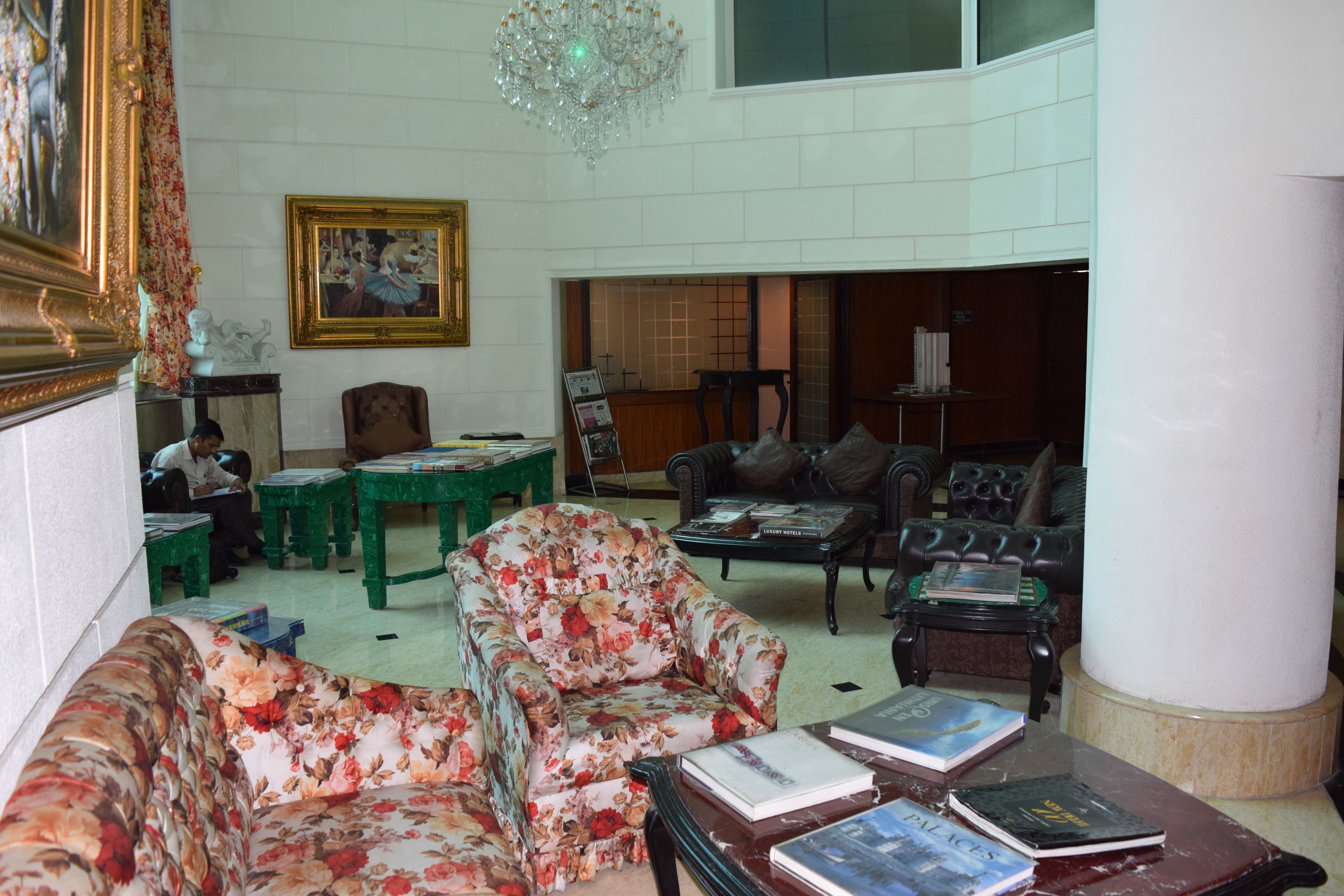 A spacious living room with a high ceiling, a chandelier, and a fireplace. There are multiple seating options including a floral armchair, a sofa, and a leather couch. The room has a large window and a framed painting on the wall.