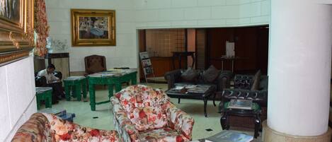 A spacious living room with a high ceiling, a chandelier, and a fireplace. There are multiple seating options including a floral armchair, a sofa, and a leather couch. The room has a large window and a framed painting on the wall.