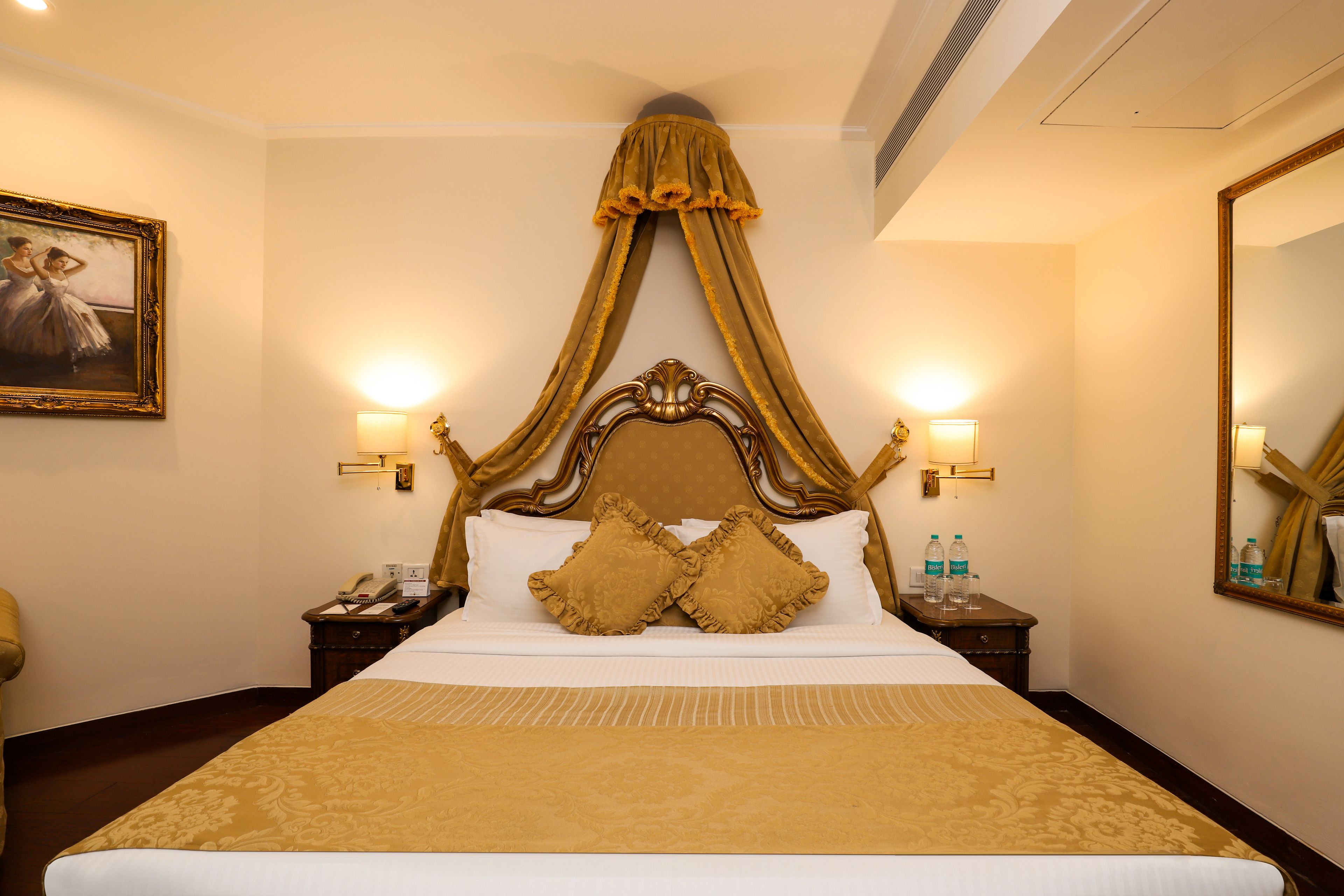 A hotel room with a large bed, a canopy bed runner, two bedside lamps, a nightstand with a phone and water bottles, and a framed picture on the wall.