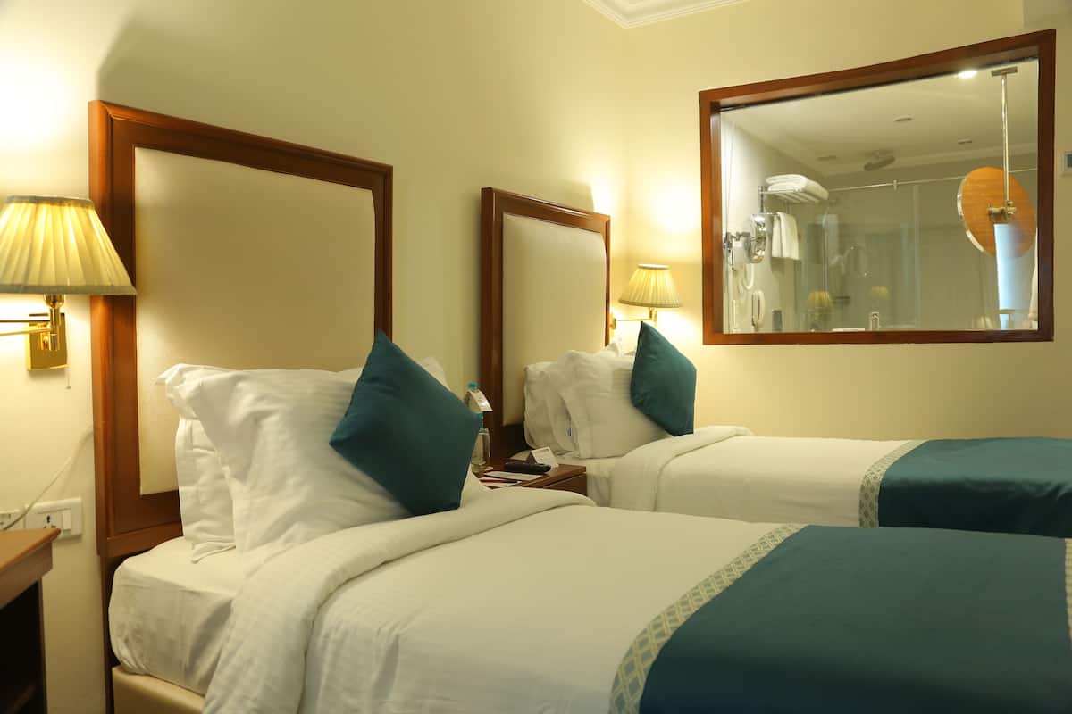 A hotel room with two beds, each with white bedding and teal pillows, a nightstand with a lamp, and a large mirror on the wall.