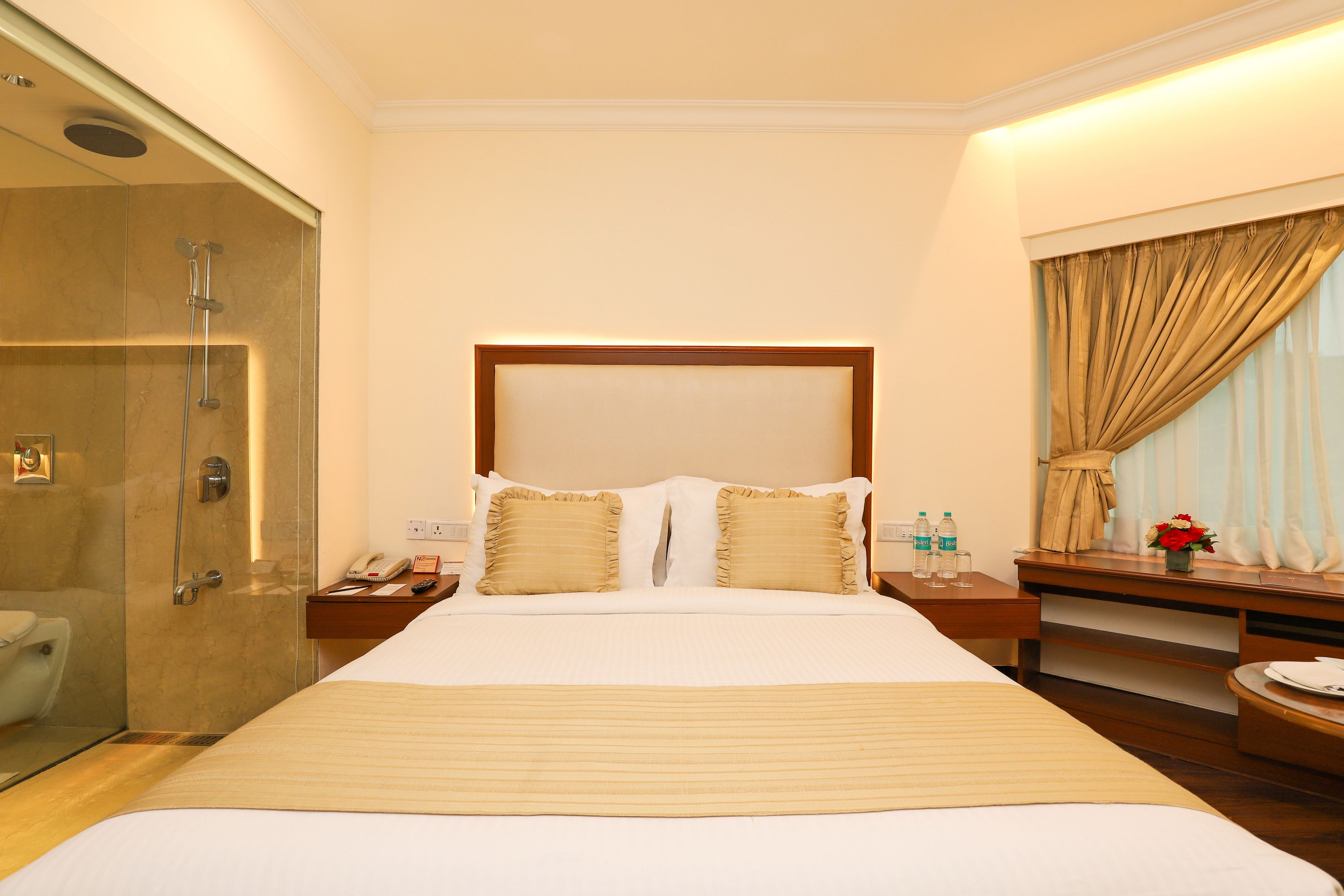 Habitación de hotel con una cama grande, una mesita de noche con lámpara, un buró con teléfono, cabina de ducha de vidrio y una ventana con cortinas.