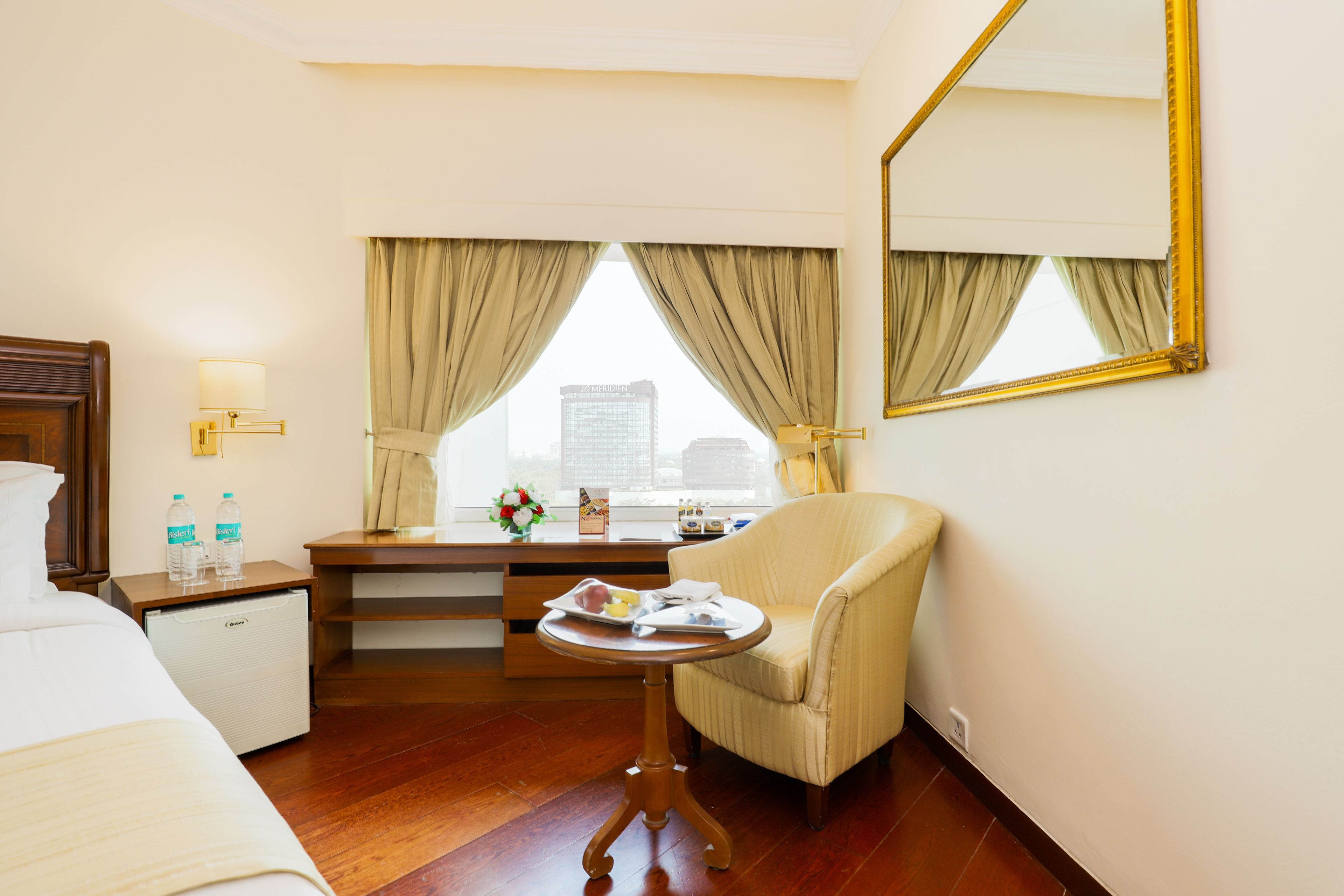 A hotel room with a bed, a desk with a lamp, a chair, a small table with a tray, and a large mirror on the wall.