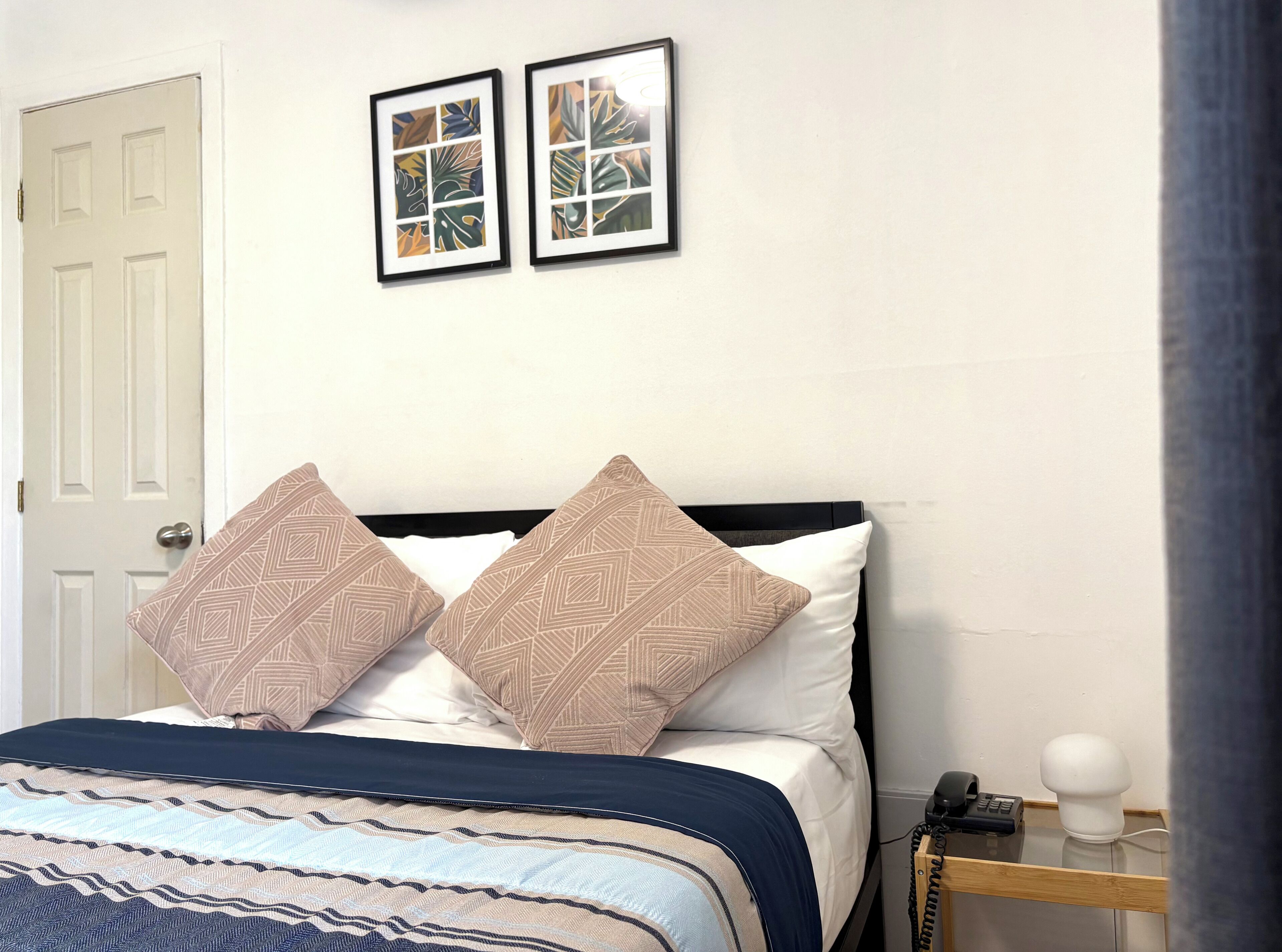 Ein ordentlich bezogenes Bett mit gemusterten Kissen, ein Nachttisch mit Telefon und Lampe sowie zwei gerahmte Bilder an der Wand.