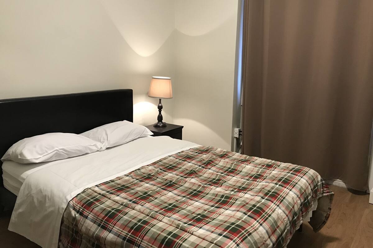 Una cama con ropa blanca, una manta de cuadros, una mesita de noche negra con una lámpara y una cortina marrón.