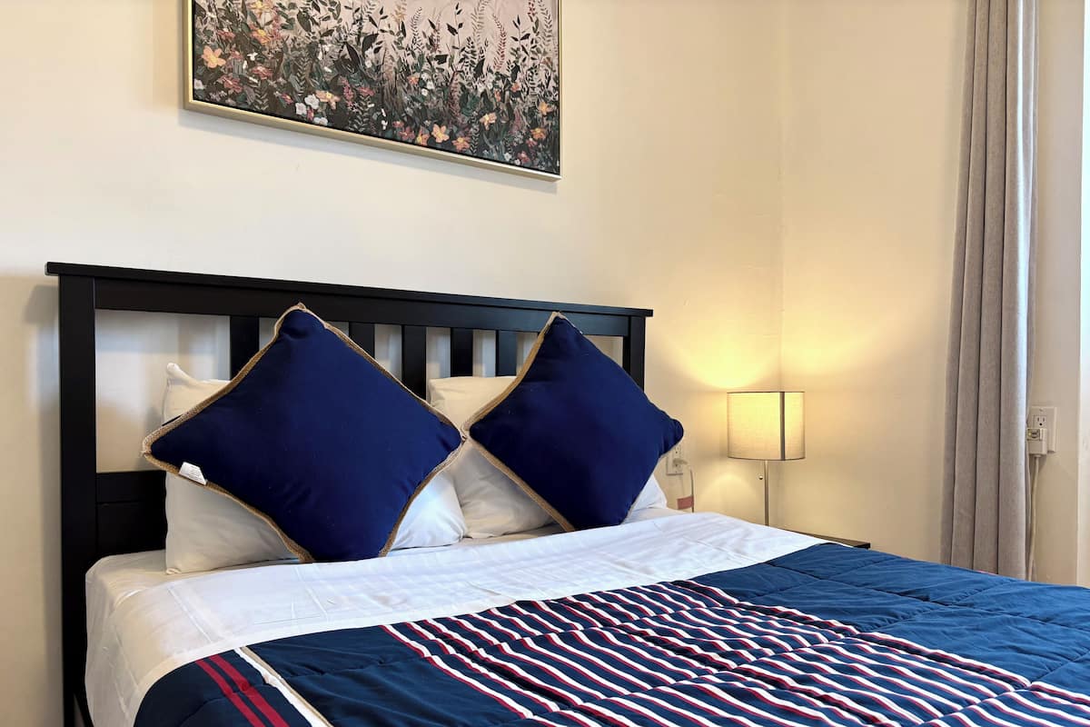 Habitación de hotel con una cama, dos almohadas, una lámpara de noche y una obra de arte floral en la pared.