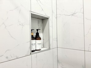 Une salle de bain avec des carreaux de marbre blanc et une étagère contenant deux bouteilles de gel douche et de shampoing.