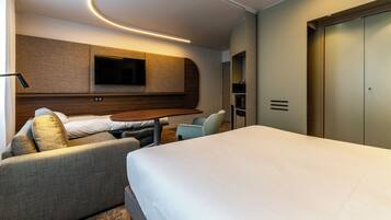A modern hotel room with a bed, a sofa, a small table, a TV mounted on the wall, and a wardrobe.