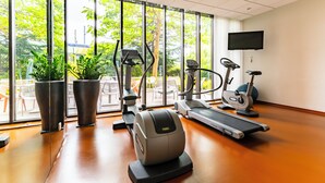 A gym with cardio machines, a TV, and large windows with a view of greenery. - Novotel Suites Reims Centre (Reims)