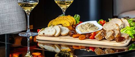 A dining table with two glasses of white wine, a wooden board with sliced bread, a bowl of dip, and assorted fruits and vegetables.