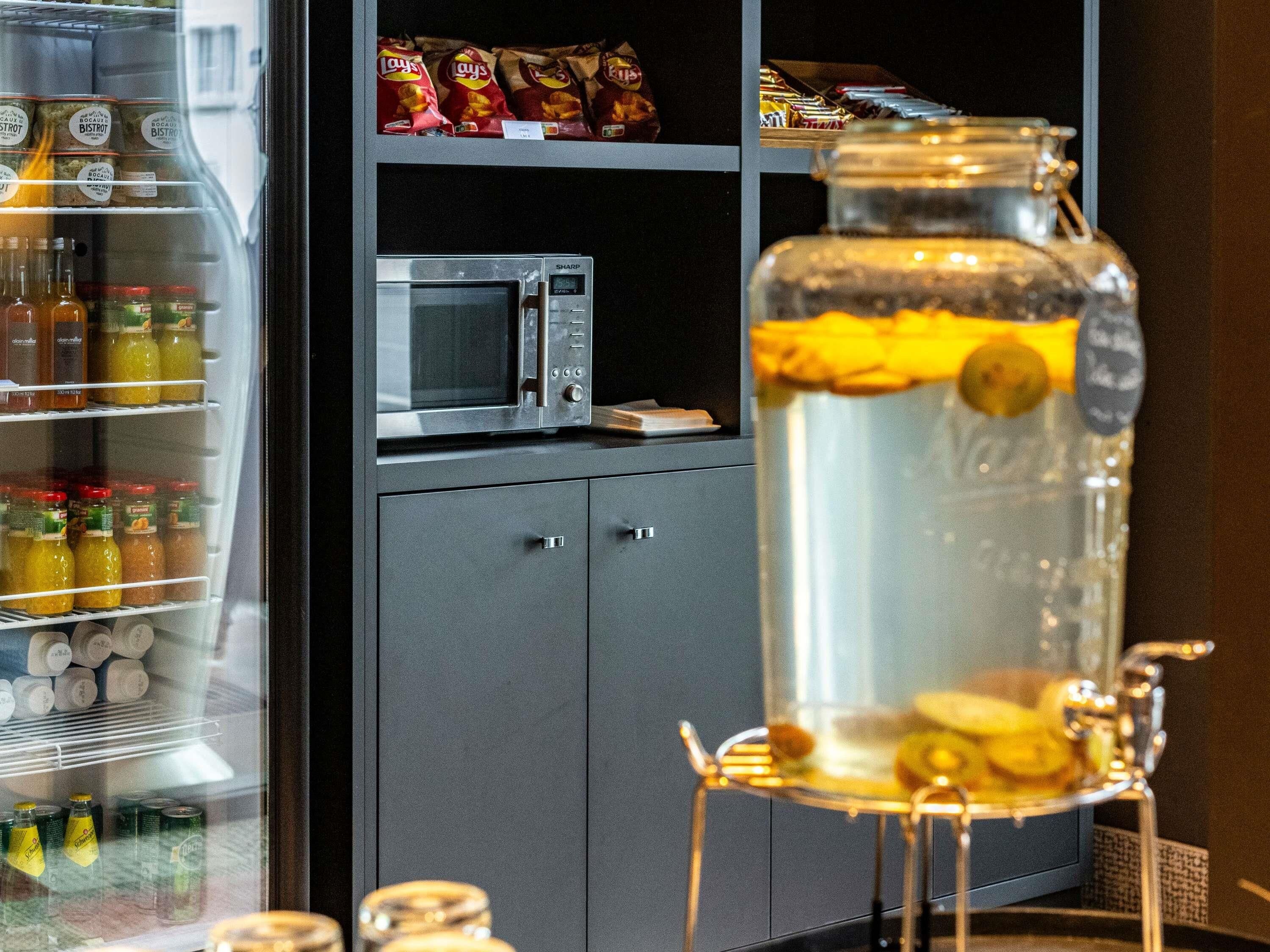 A kitchen area with a refrigerator stocked with beverages, a microwave, and a glass jar with a metal stand.