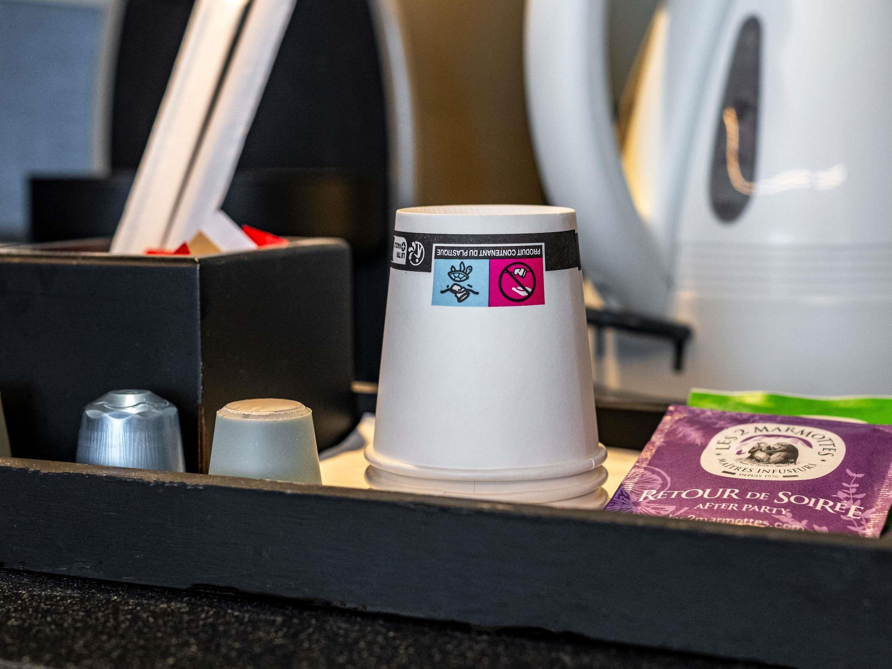 Una cafetera con vasos desechables, un pequeño cubo de basura y una bolsita de aperitivos.