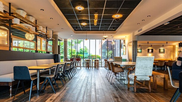 A modern restaurant interior with wooden flooring, tables, chairs, and a large window offering a view of greenery. - Novotel Suites Reims Centre (Reims)