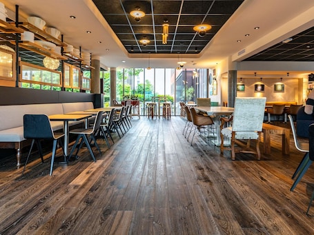 A restaurant interior with wooden flooring, modern furniture, and large windows offering a view of greenery.. Novotel Suites Reims Centre