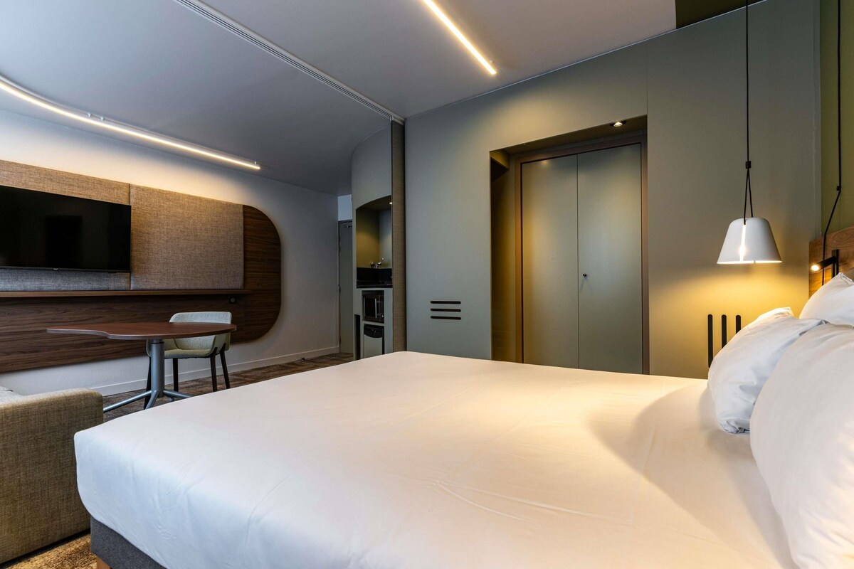 Une chambre d’hôtel moderne dotée d’un grand lit, d’un téléviseur à écran plat fixé au mur, d’une petite table avec une chaise et d’une armoire.