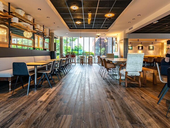 A modern restaurant interior with wooden flooring, tables, chairs, and a large window offering a view of greenery. - Novotel Suites Reims Centre (Reims)