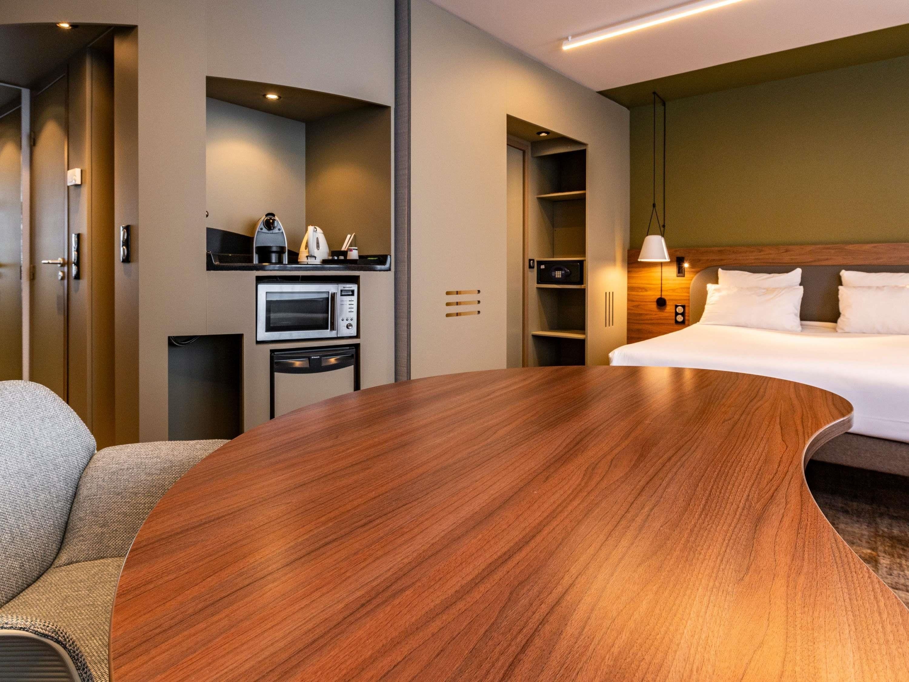 A hotel room with a curved wooden table, a bed with white linens, a microwave, a coffee maker, and a built-in wardrobe.
