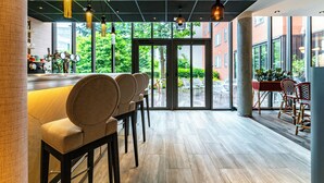 A modern bar area with a long counter, several cushioned bar stools, and pendant lighting. Large windows provide views of the outdoors. - Novotel Suites Reims Centre (Reims)
