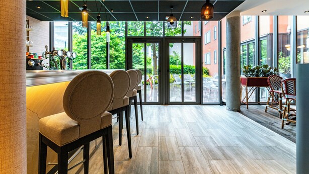 Un área de bar moderna con una barra alargada, varios taburetes acolchados e iluminación colgante. Amplias ventanas ofrecen vistas al exterior.