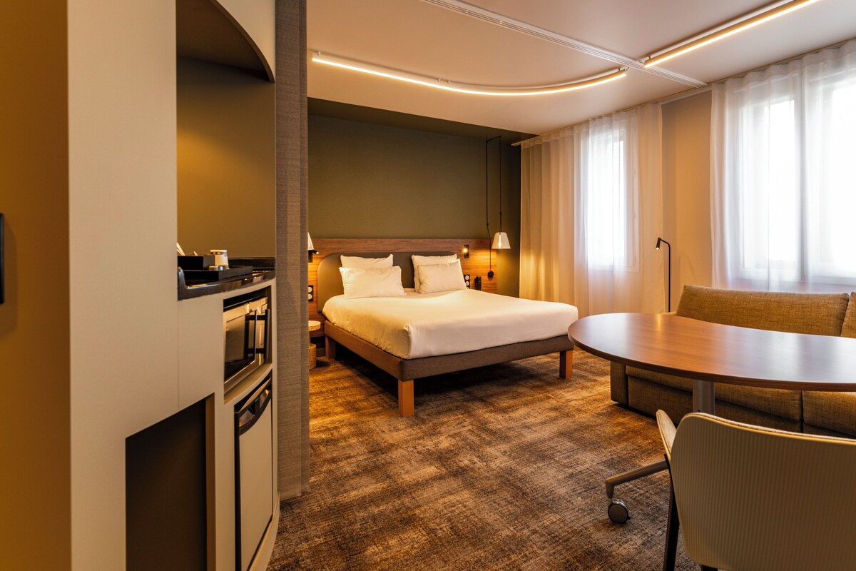 A hotel room with a bed, a sofa, a table, and a desk with a lamp.