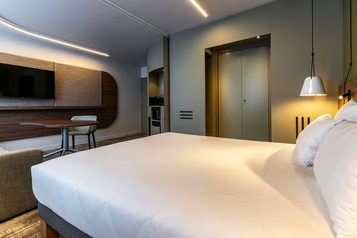 Una habitación de hotel moderna con una cama grande, un televisor de pantalla plana en la pared, una mesita con una silla y un armario.