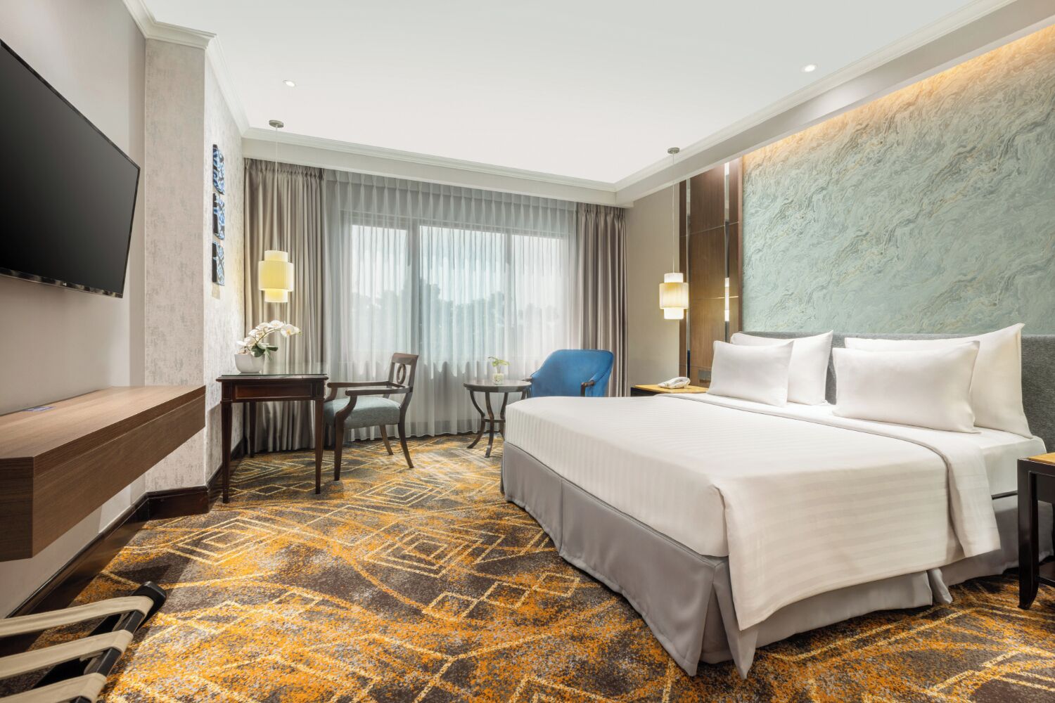 A hotel room with a large bed, a desk with a chair, a TV mounted on the wall, and a patterned carpet.