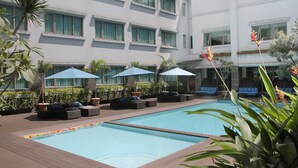 Ein Hotelpoolbereich mit mehreren Liegestühlen und Sonnenschirmen, umgeben von Grünflächen und einem Gebäude mit großen Fenstern.