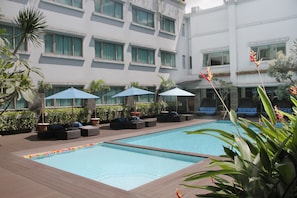 Ein Hotelpoolbereich mit mehreren Liegestühlen und Sonnenschirmen, umgeben von Grünflächen und einem Gebäude mit großen Fenstern.