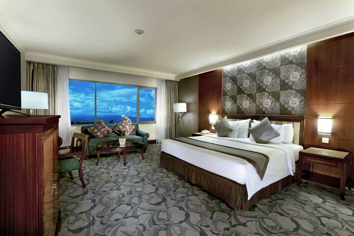 Habitación de hotel con una cama grande, un sofá, un televisor y vistas del cielo a través de un ventanal.