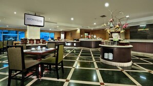 Een ruime hotel lobby met een geruit vloertapijt, een bar in het midden en een televisiescherm waarop de naam van het hotel staat.
