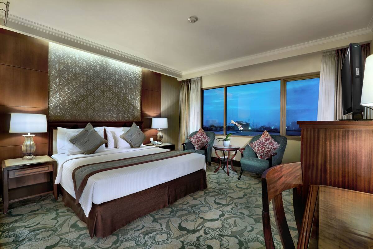 Habitación de hotel con una cama grande, dos mesitas de noche, un escritorio con silla, vista a la ciudad y un ventanal amplio.