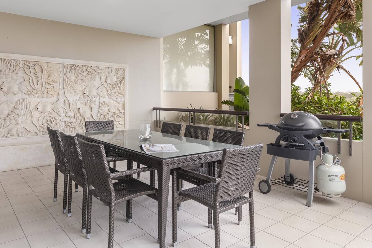 Un espace repas comprenant une table en verre, des chaises en osier, un barbecue et une bouteille de gaz.