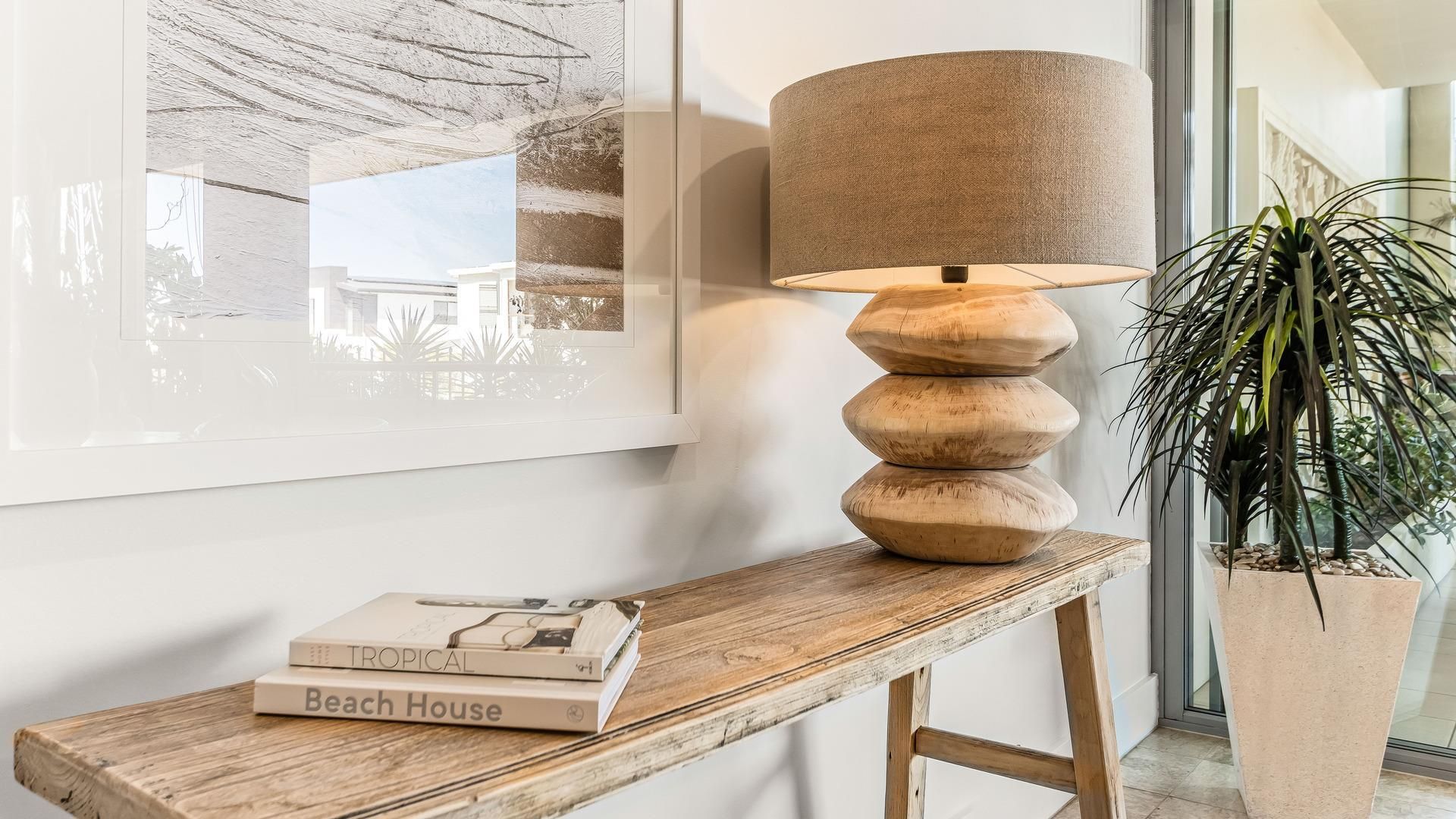 Ett träbord med en lampa och böcker, en inramad tavla på väggen och en krukväxt.
