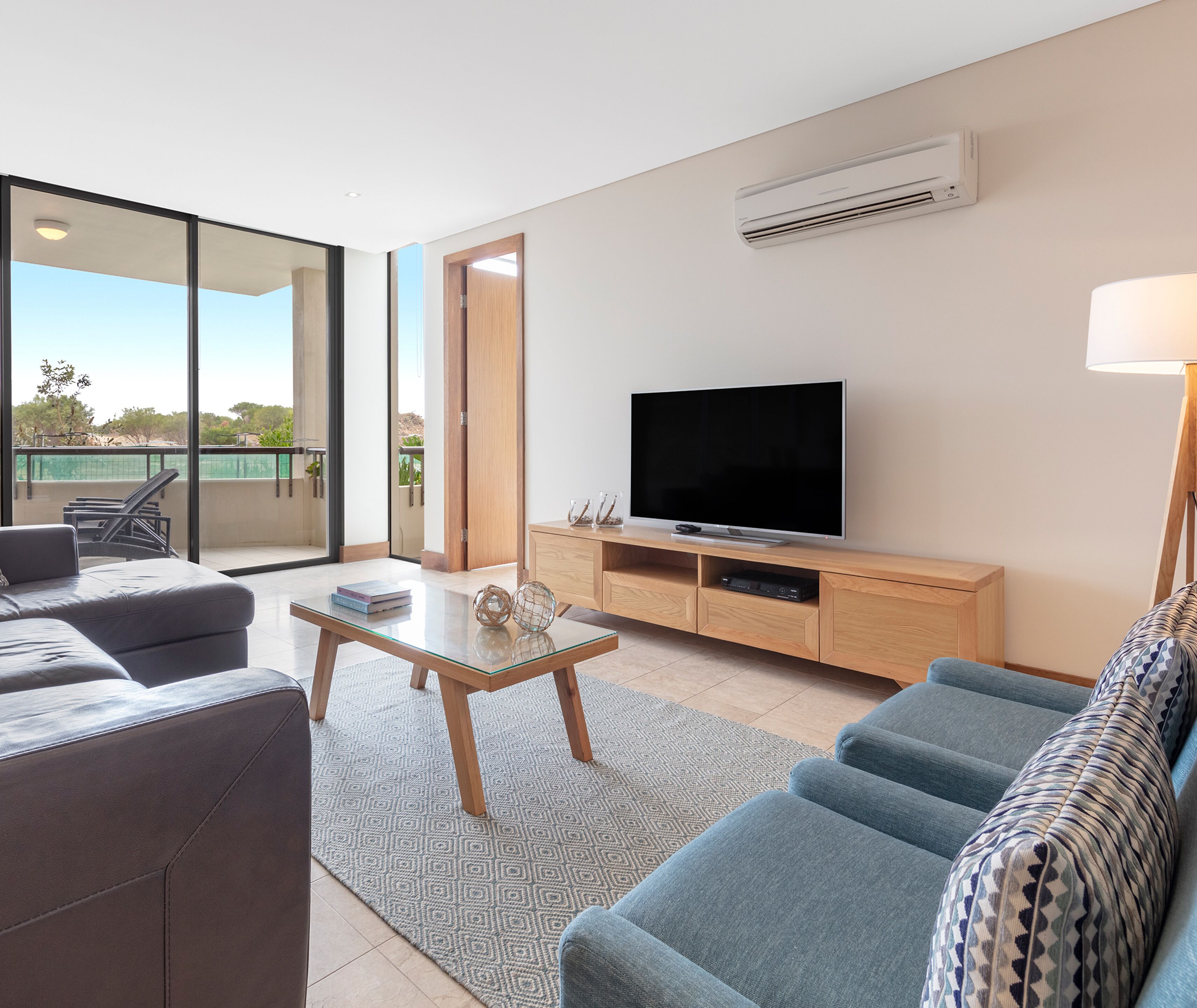 A modern living room with a large flat-screen TV, comfortable seating, a glass coffee table, and a view of the outdoors.
