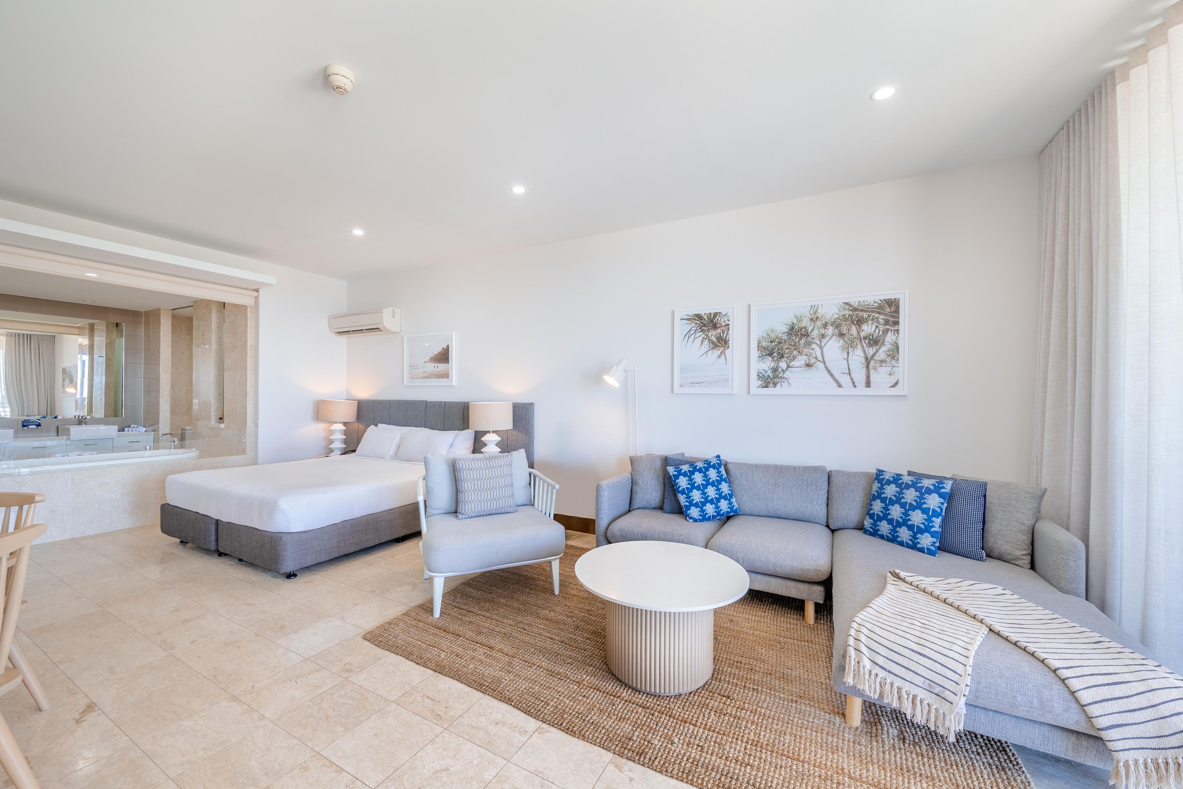 A modern living room with a sofa, armchair, and a round coffee table. There is a bed with a headboard and a bathroom visible in the background.