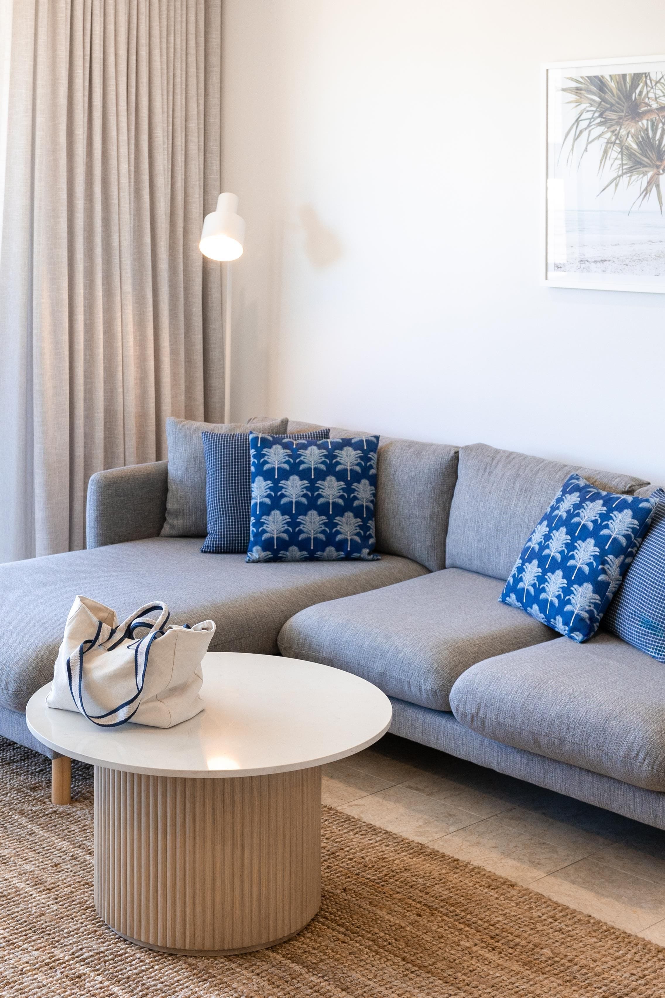 A modern living room with a grey sofa, two patterned pillows, a white coffee table, and a beige bag.
