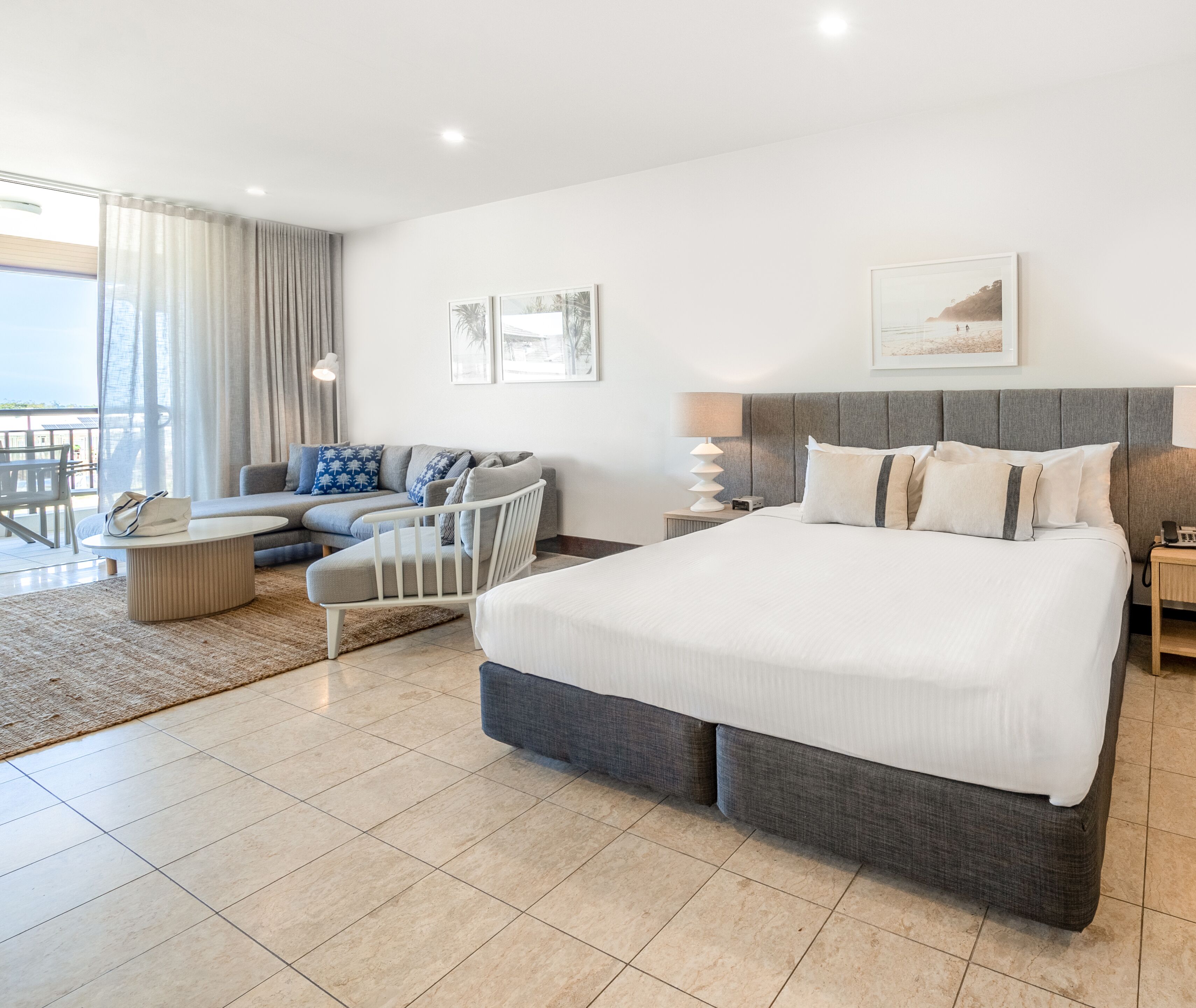 A modern hotel room with a large bed, a sofa, a chair, and a balcony with curtains.