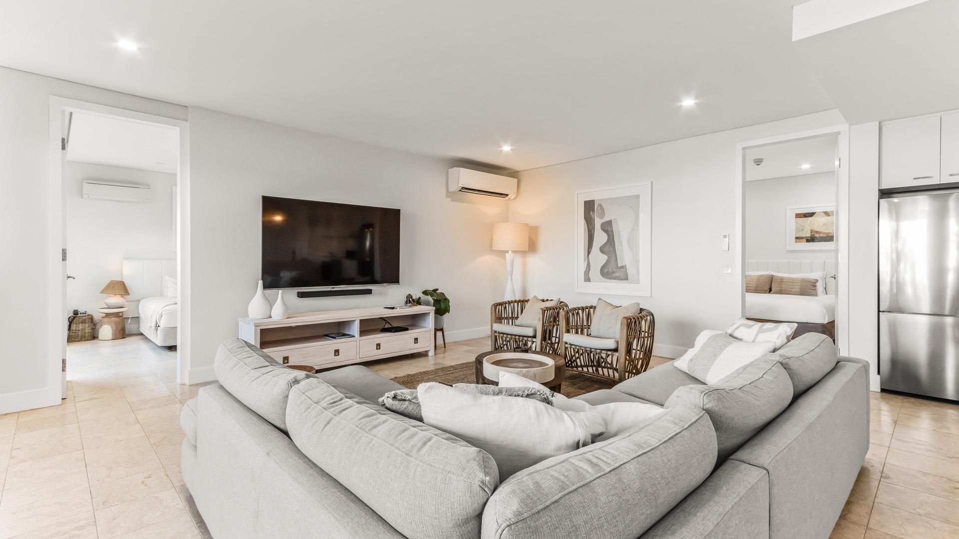 A modern living room with a grey sofa, a flat-screen TV, a white entertainment unit, wicker chairs, and a kitchen area with a stainless steel refrigerator.