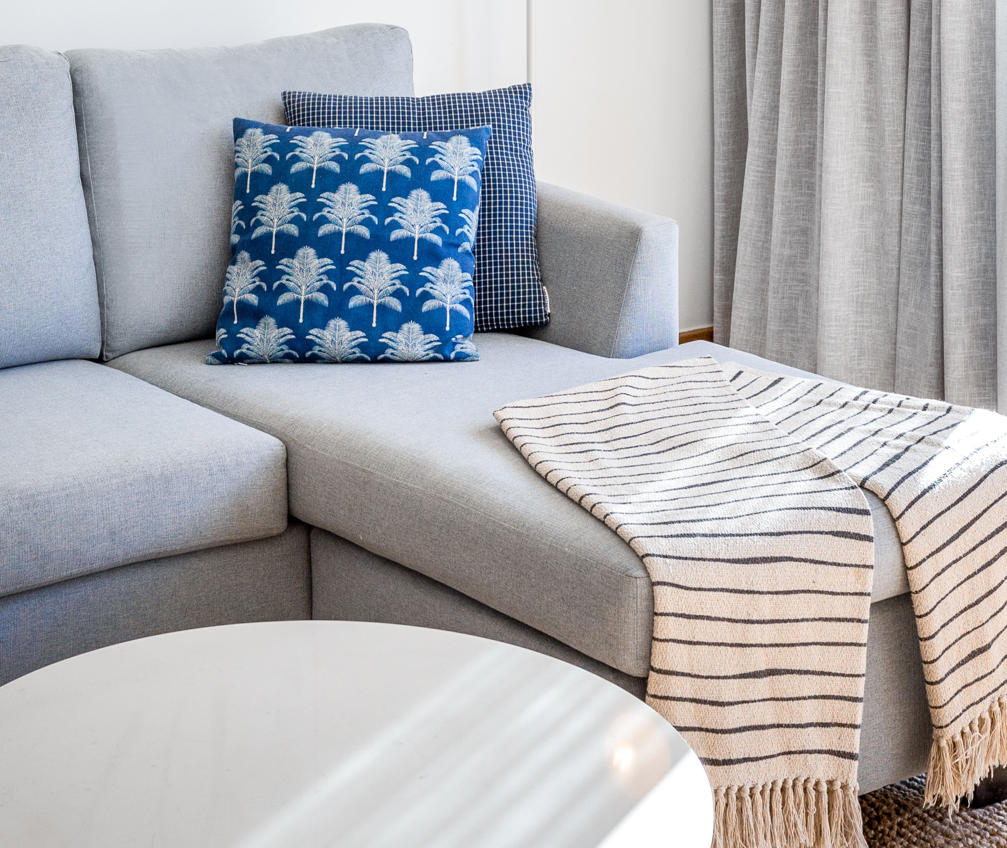 A grey sofa with a blue and white patterned pillow, a striped throw blanket, and a white round table.