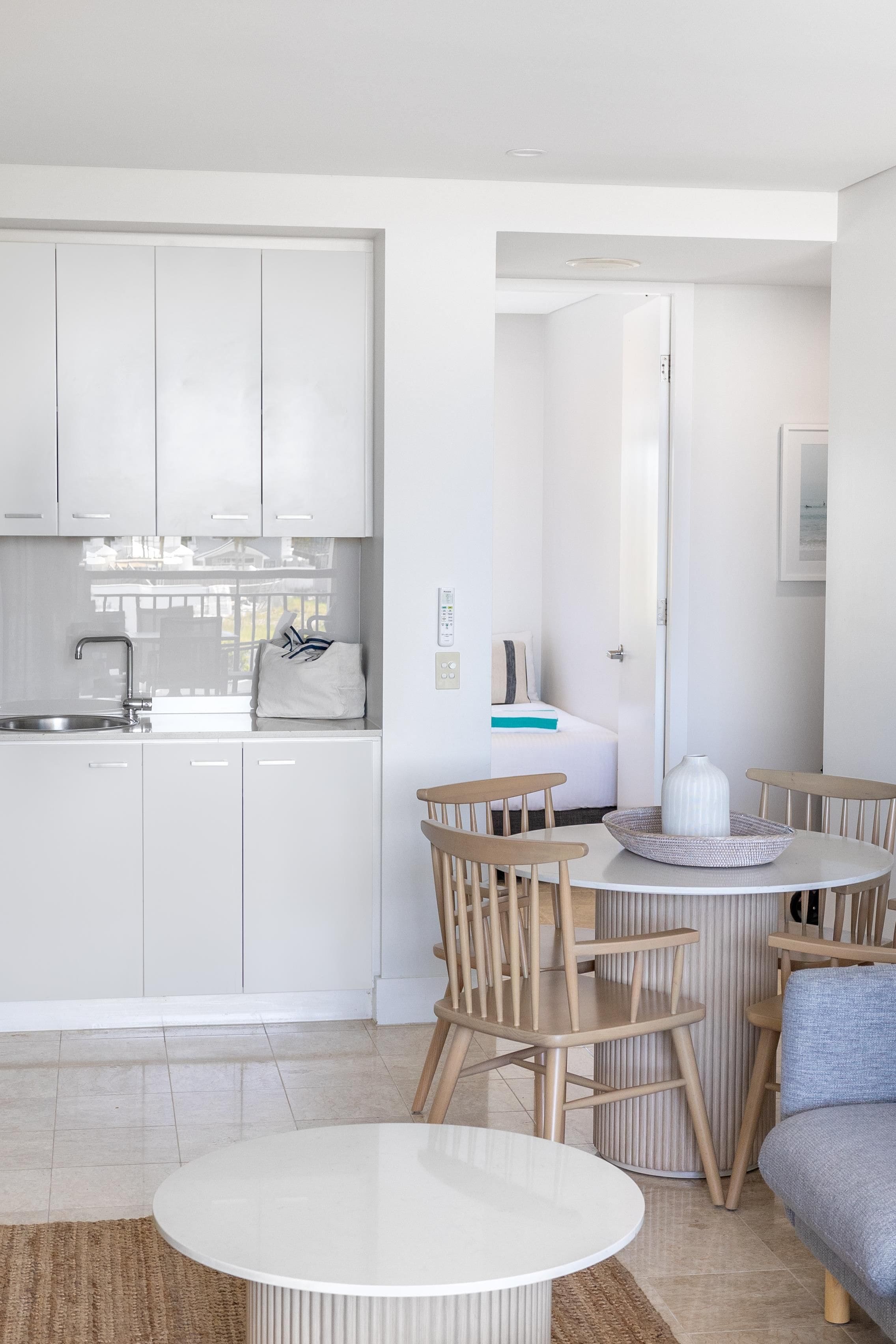 Una cocina moderna con armarios blancos, un comedor con una mesa redonda y sillas de madera, y vista al mar a través de una puerta abierta.