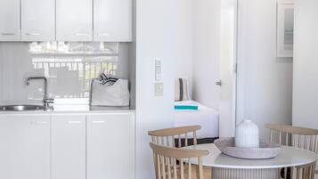 A modern kitchen with white cabinets, a dining area with a round table and wooden chairs, and a view of the sea through an open door.