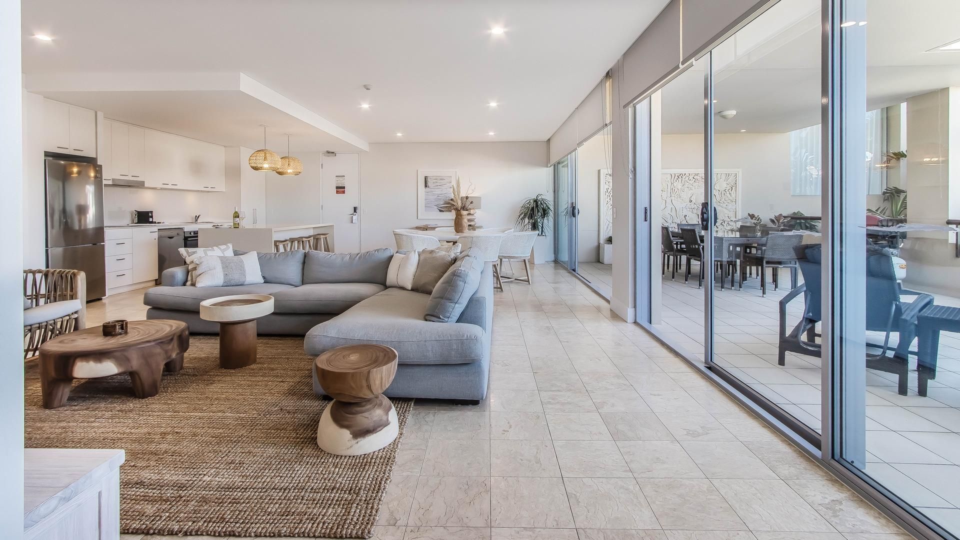 A modern living room with a grey sofa, wooden coffee table, and a dining area with outdoor seating.