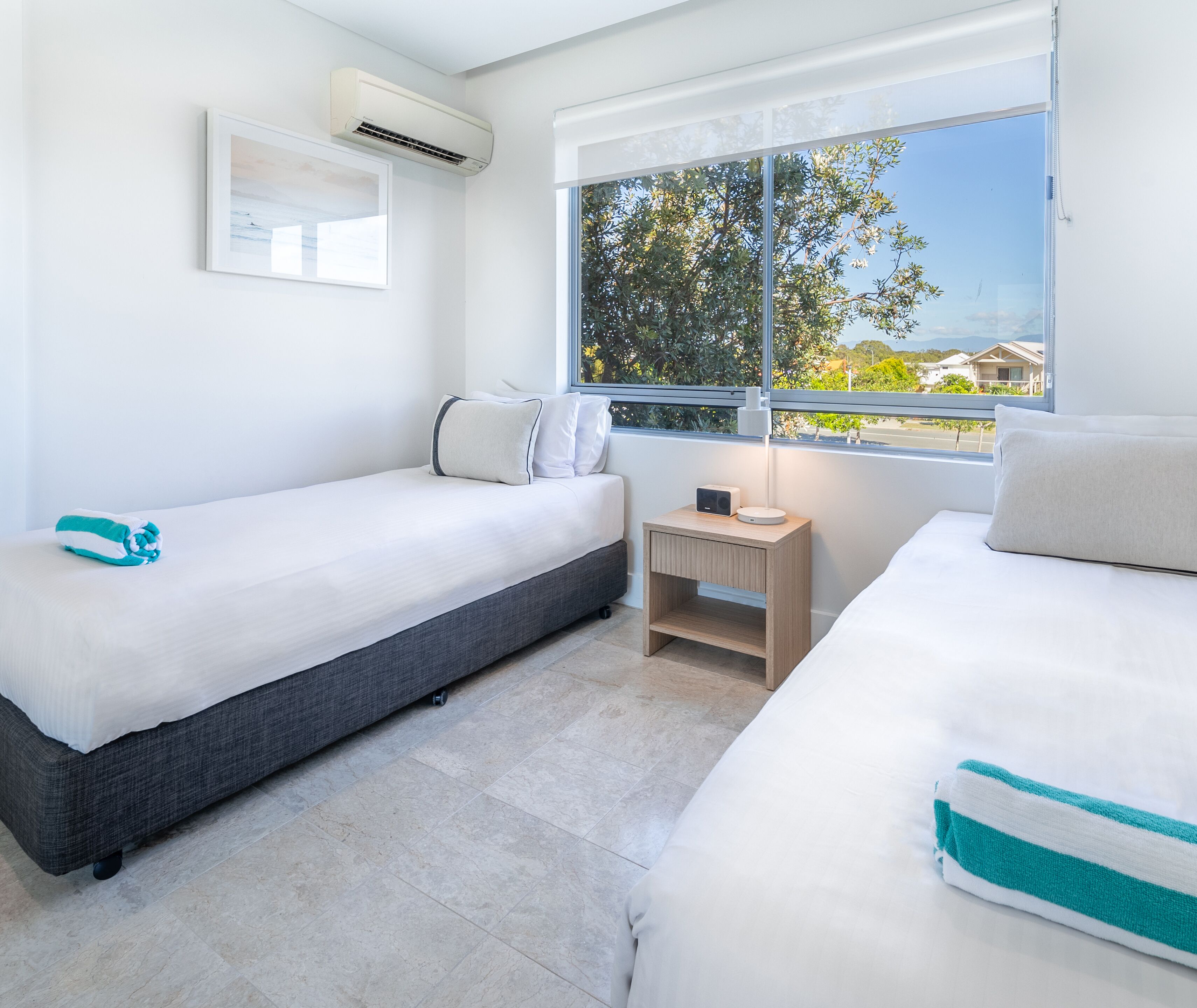 Quarto de hotel moderno com duas camas, uma mesinha de cabeceira com abajur e uma janela com vista para árvores e edifícios.