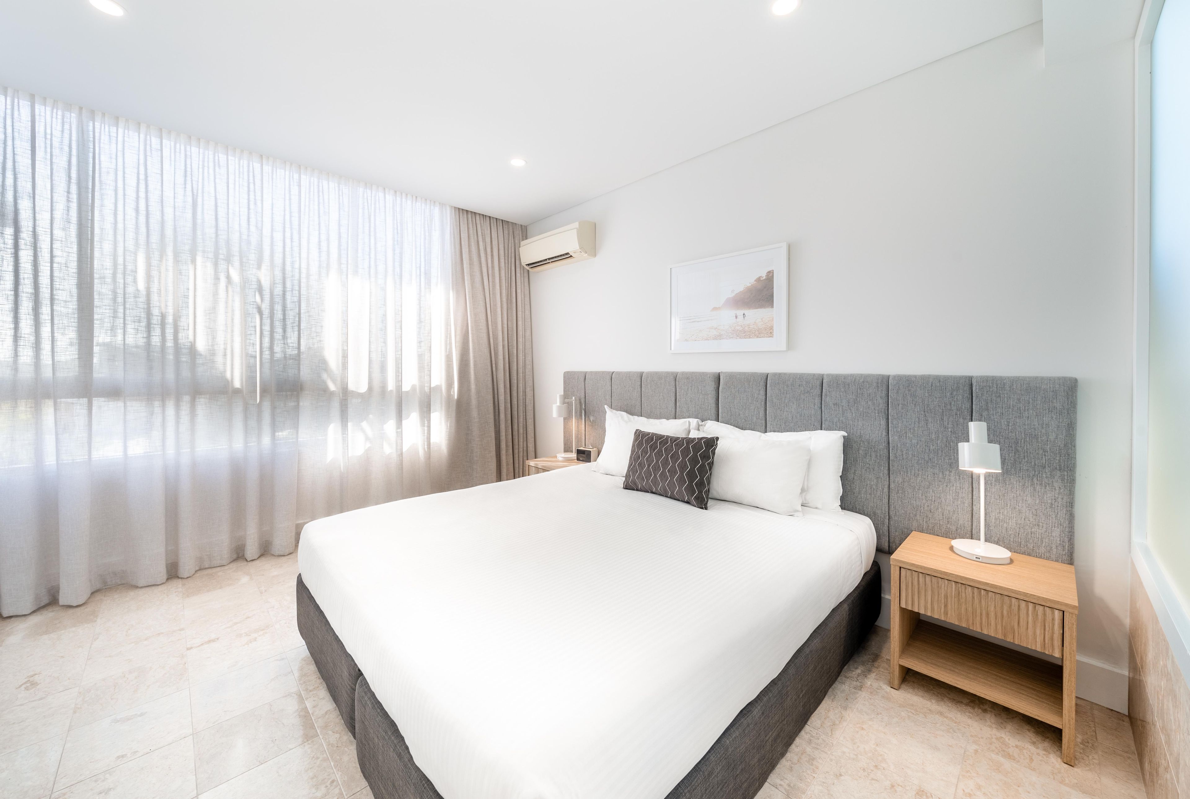 A modern hotel room with a large bed, a nightstand, a lamp, and a view of the outdoors through sheer curtains.