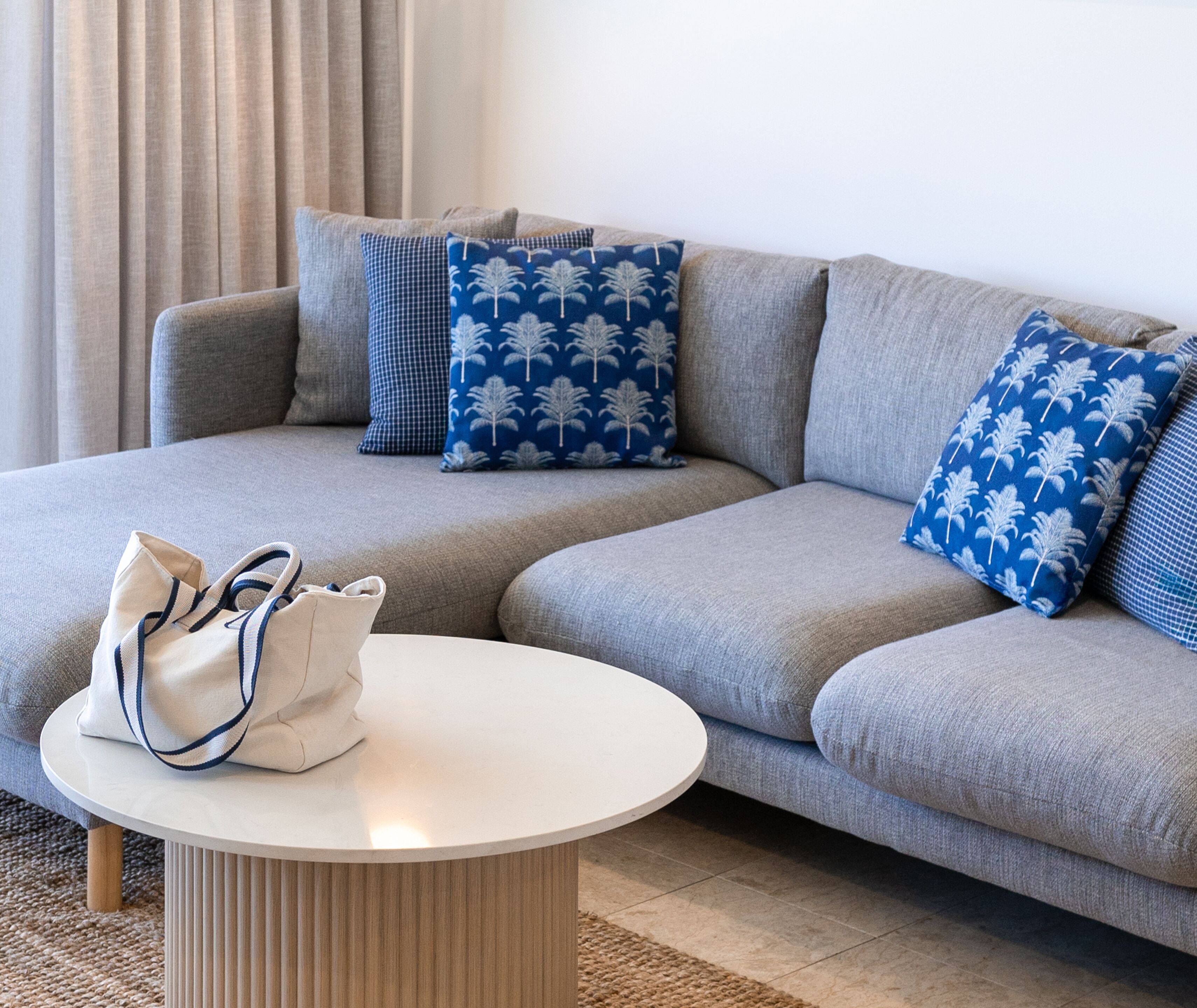 A grey sofa with blue and checkered cushions, a white round coffee table, and a beige handbag.