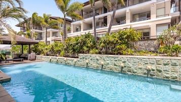 A swimming pool with a stone wall, surrounded by palm trees and a multi-story building with balconies.